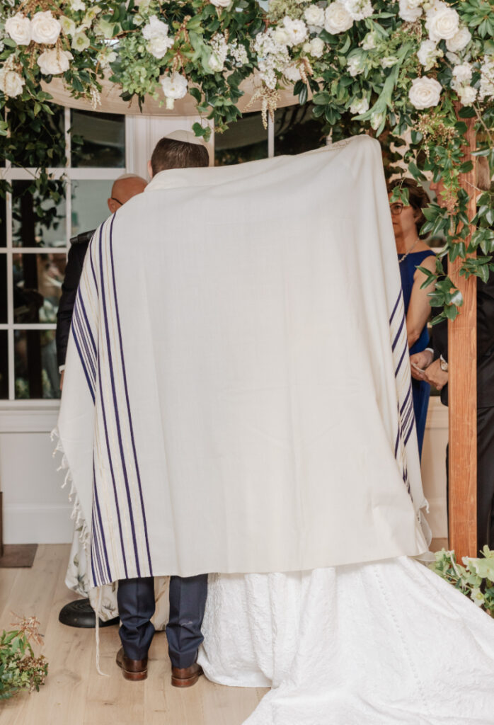 Couple standing beneath prayer shawl during ceremony, meaningful tradition surrounded by family and florals