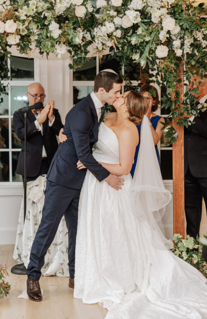 Couple sharing first kiss under floral canopy, joyful ceremony moment surrounded by applause